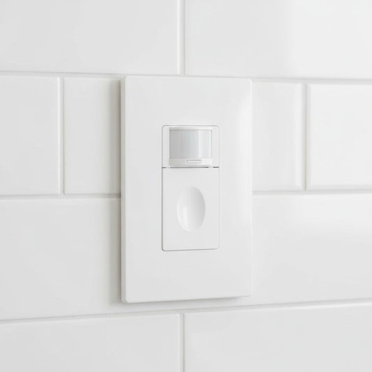 A rectangular white light switch features a semi-transparent sensor lens and a flush manual button mounted on a tiled wall. Soft daylight highlights the clean edges of the faceplate and the modern sensor window.