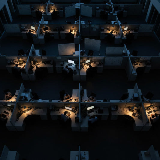 A high-angle wide shot looks down into a maze of gray fabric-wrapped cubicles in a modern office at twilight. Desk lamps cast warm pools of light while the general overhead lighting is turned off.