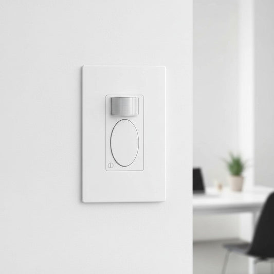 A rectangular white motion sensor switch is mounted flush against a painted wall in an office setting. The central lens detects movement to control room lighting automatically.